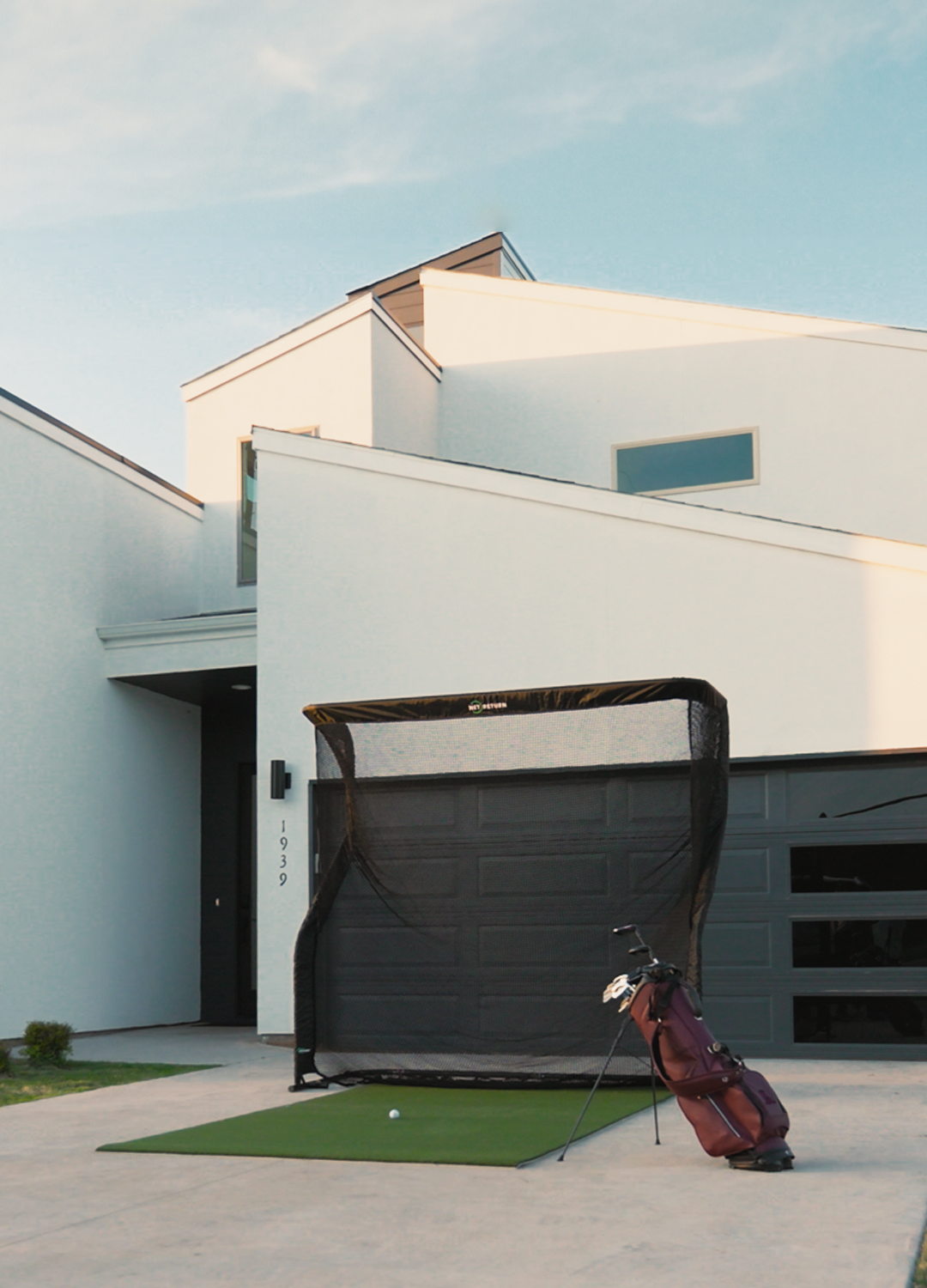Modern white house with angular design, featuring a golf net setup in front of a garage. A golf bag with clubs is on the driveway's artificial turf.
