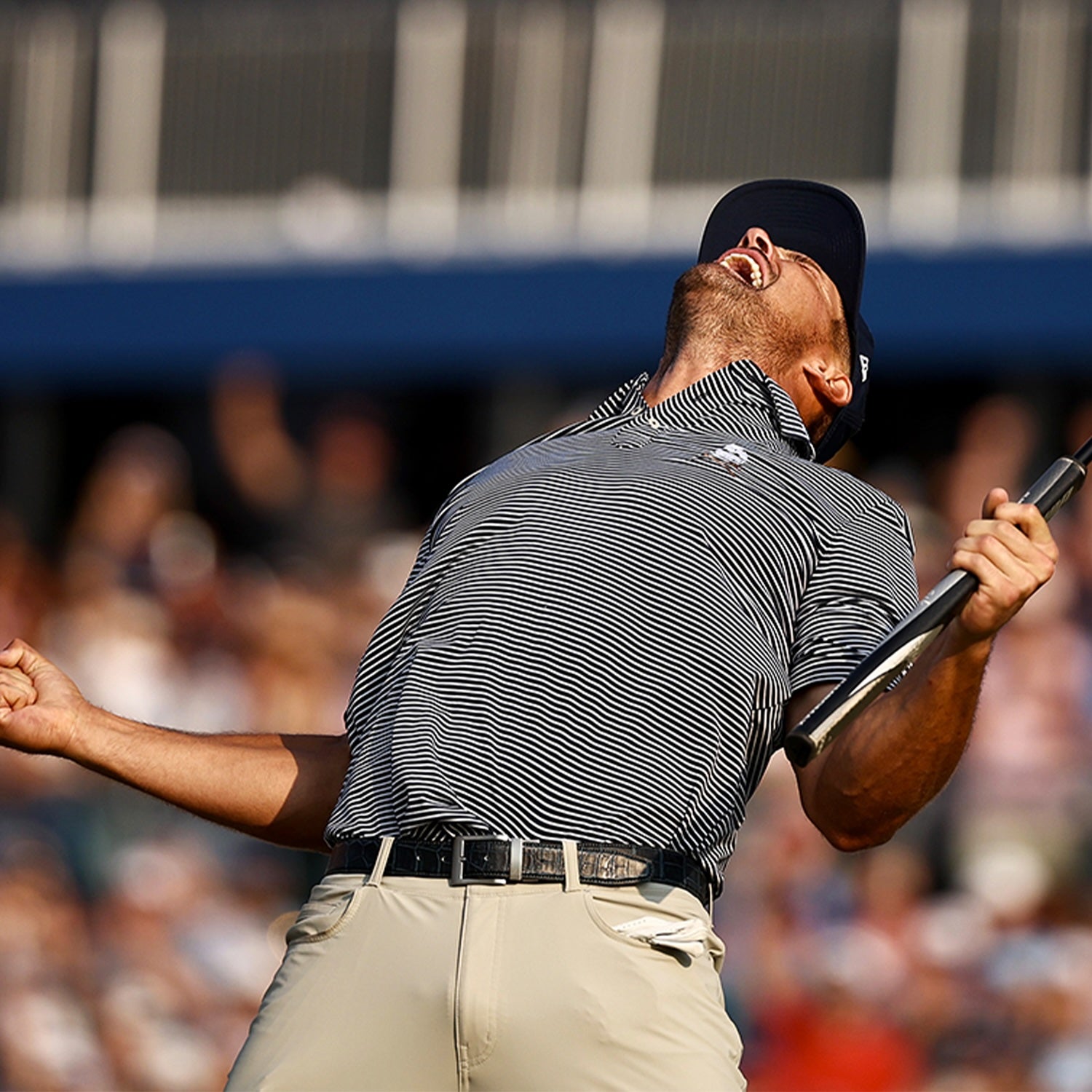 Golfer in a striped shirt and cap celebrates passionately with a putter raised, against a blurred background of spectators.