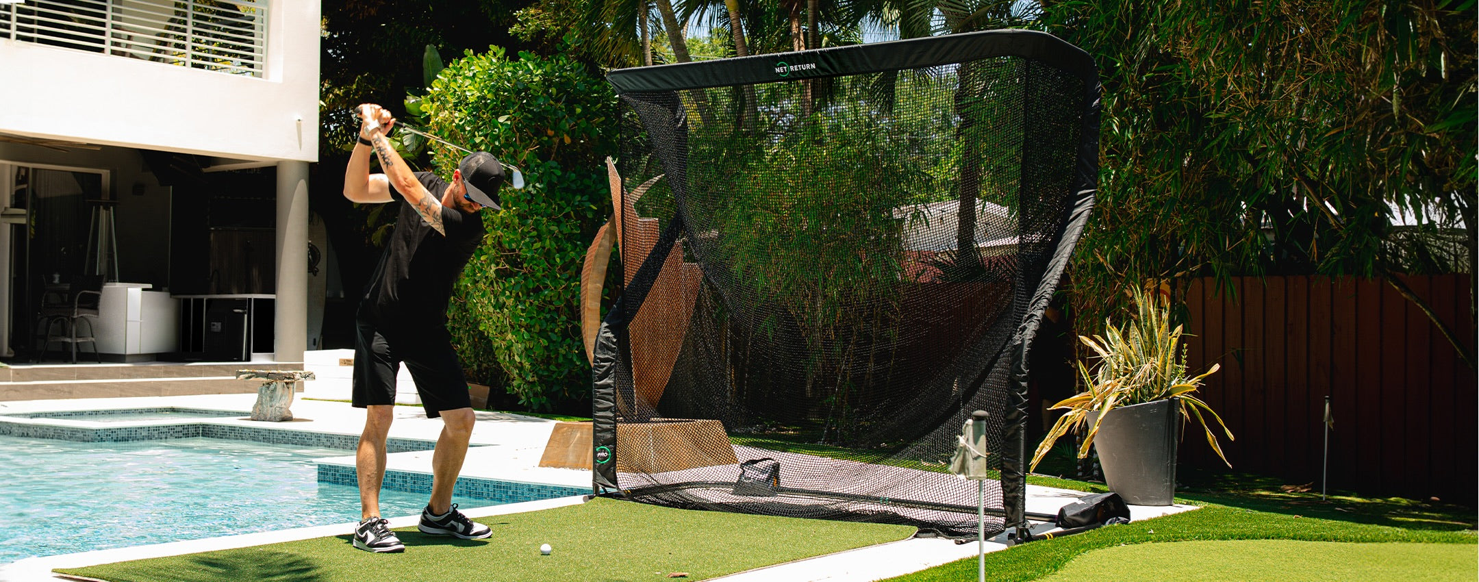 A person in a black outfit practices golf swings on a green mat near a pool, using a net for capturing golf balls.