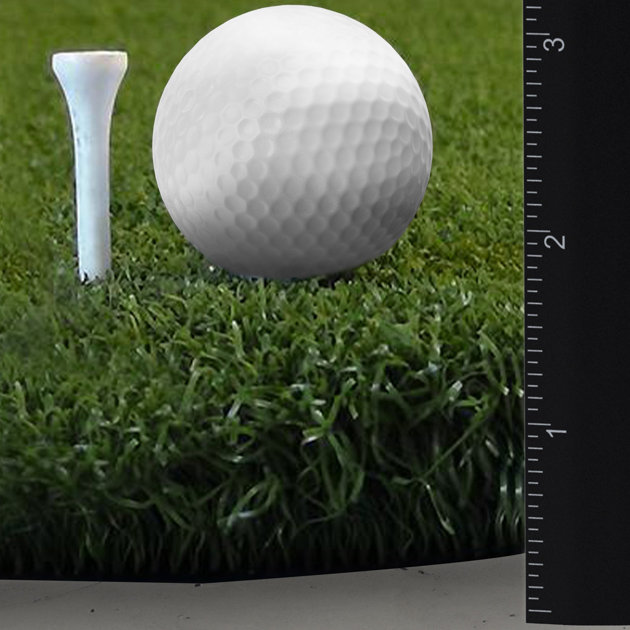 A golf ball rests on grass next to a tee, with a black ruler showing measurements in inches and centimeters.