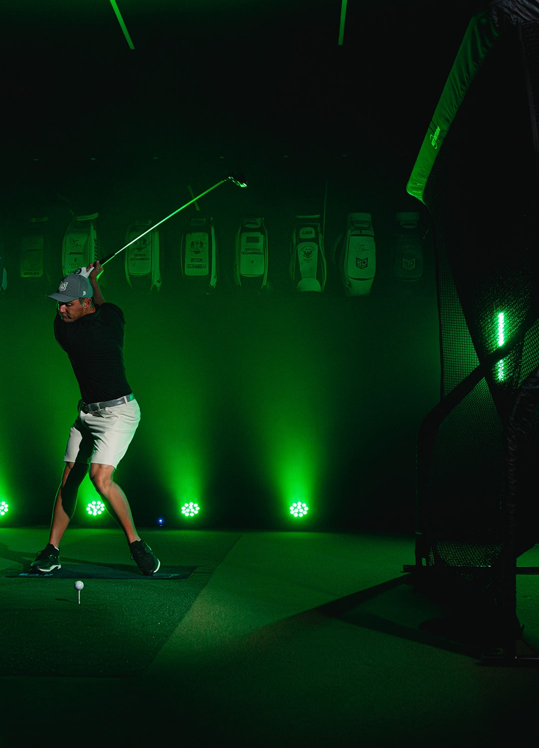 A golfer in mid-swing practices indoors with green lighting and golf bags displayed in the background.