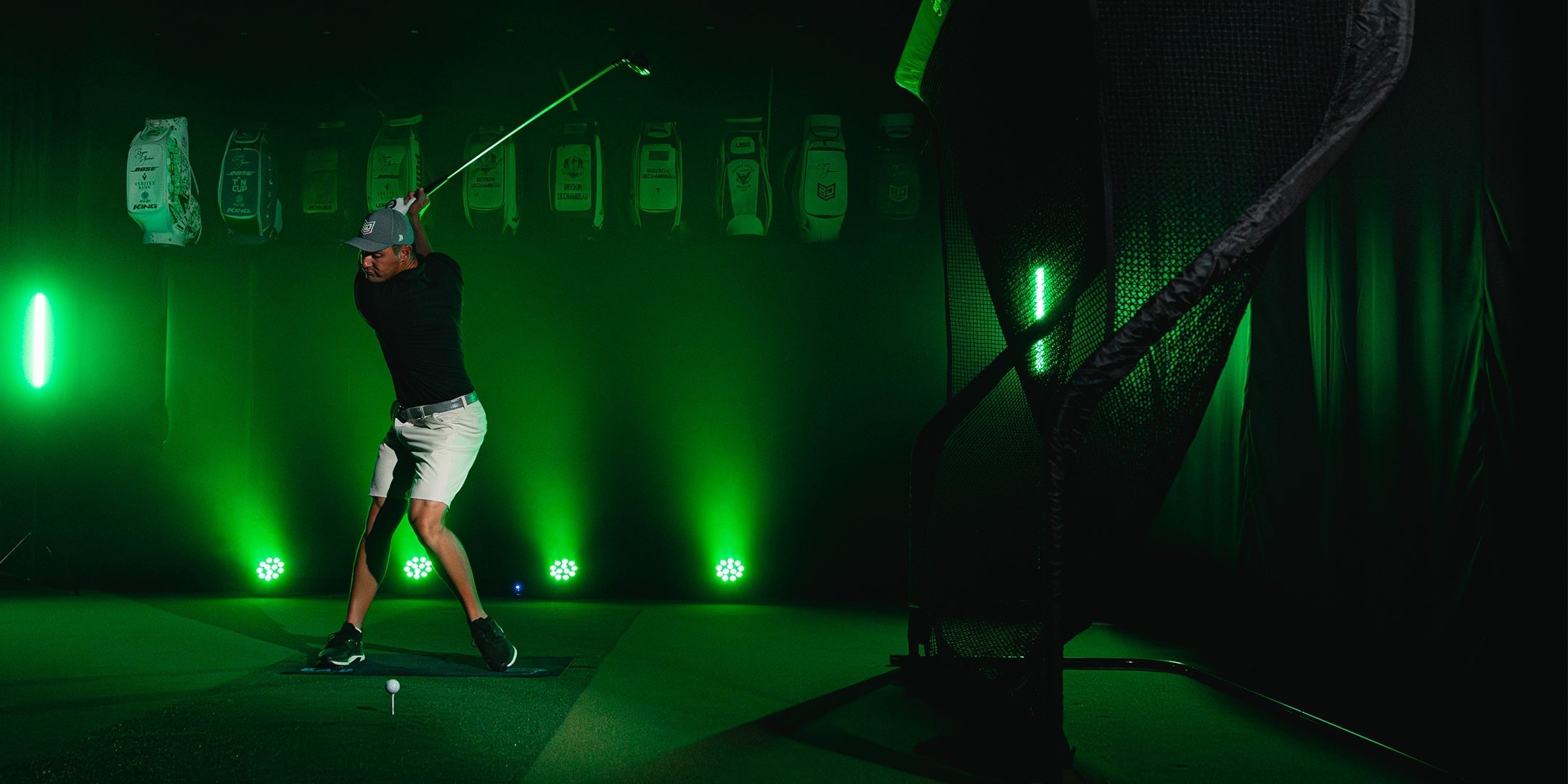 A person wearing a cap and shorts swings a golf club indoors, aiming at a net. Bright green lights illuminate the room, with golf bags visible in the background.