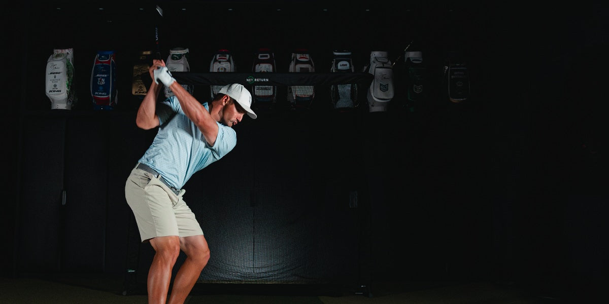 A golfer in a blue shirt and beige shorts prepares to swing indoors, with a row of golf bags displayed in the background.