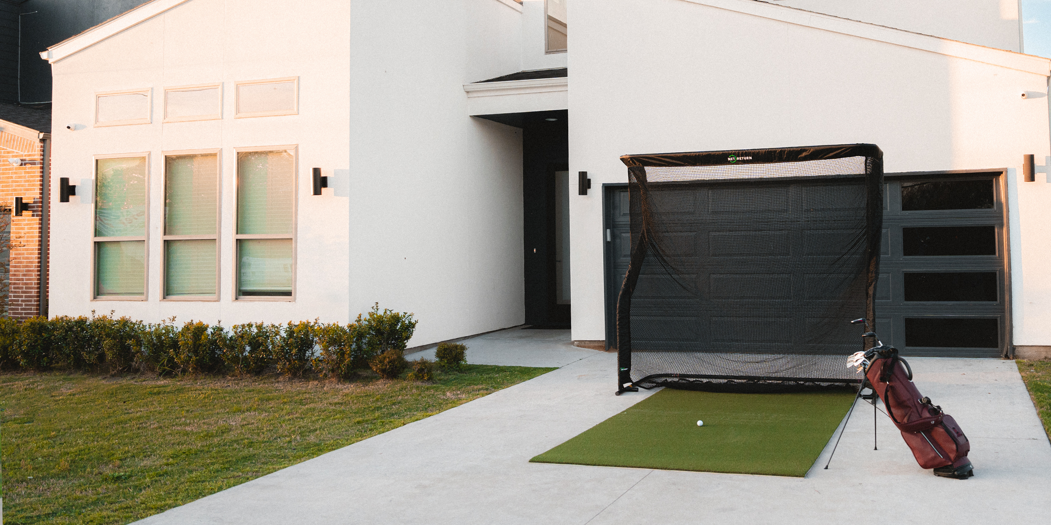 A golf practice setup with a net, a mat, and a golf bag is placed on the driveway in front of a modern white house.