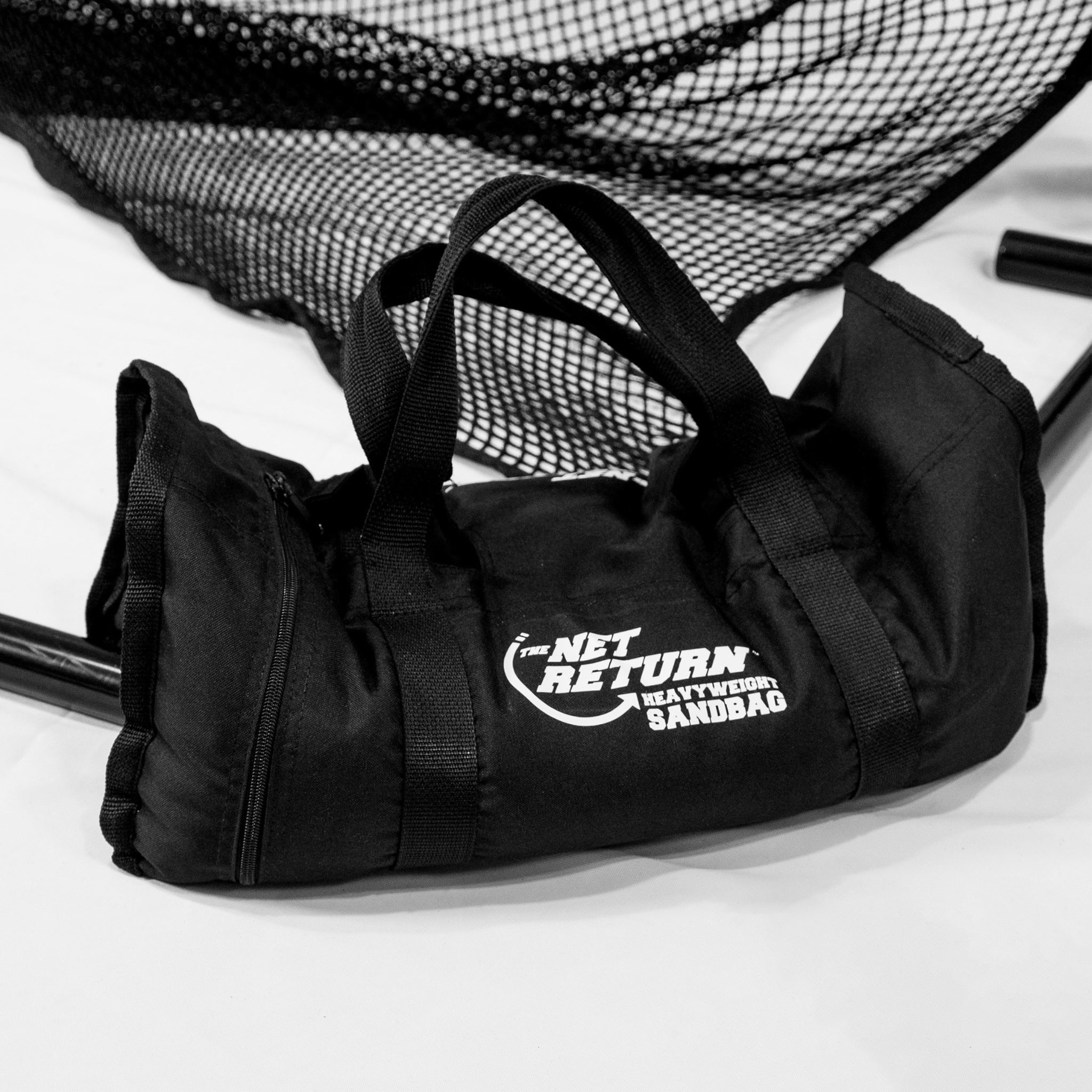 A black duffel bag labeled "The Net Return Heavyweight Sandbag" placed on a white surface with netting and metal poles in the background.