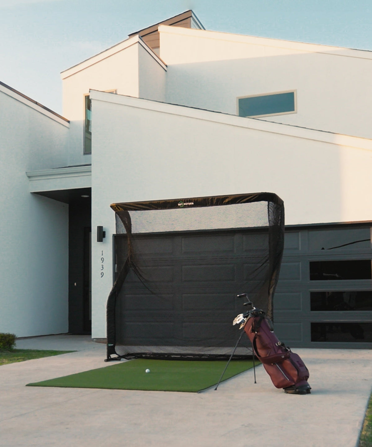 Golf practice setup with a net and mat on a driveway in front of a modern house. A golf bag with clubs is positioned to the side.