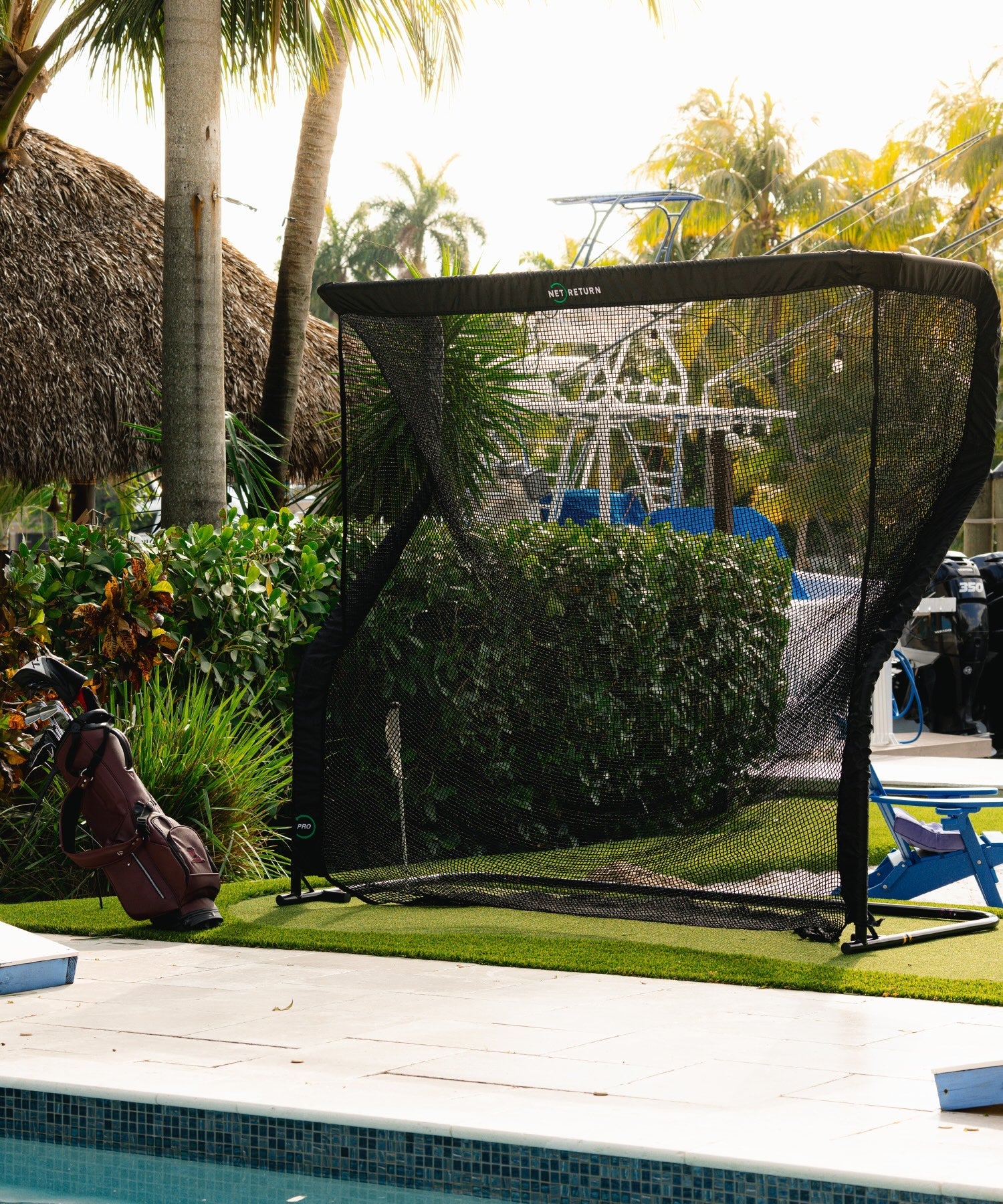 A golf practice net set up by a poolside, with a golf bag nearby. Tropical plants and a boat are in the background.