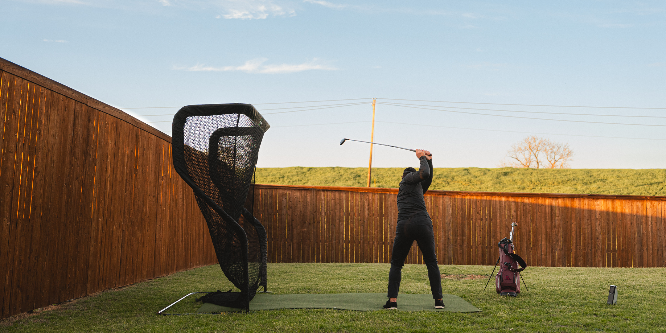 Person practicing golf swing in a backyard with a net, golf bag, and insulated tumbler nearby.